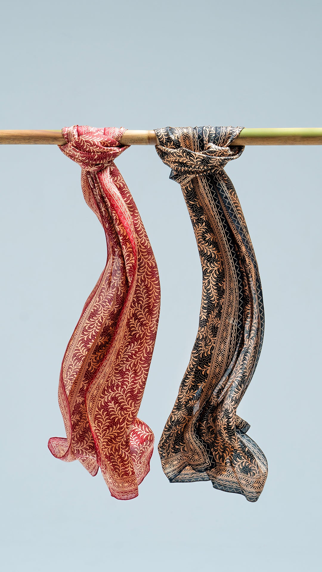 Two daun asem scarves hanging on a rod against a light blue background