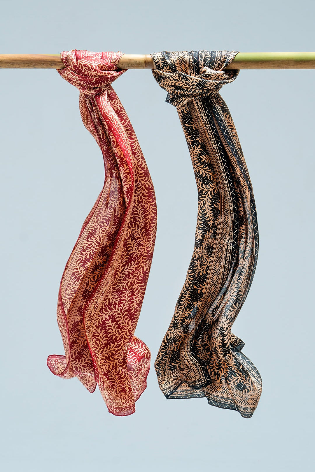 Two daun asem scarves hanging on a rod against a light blue background